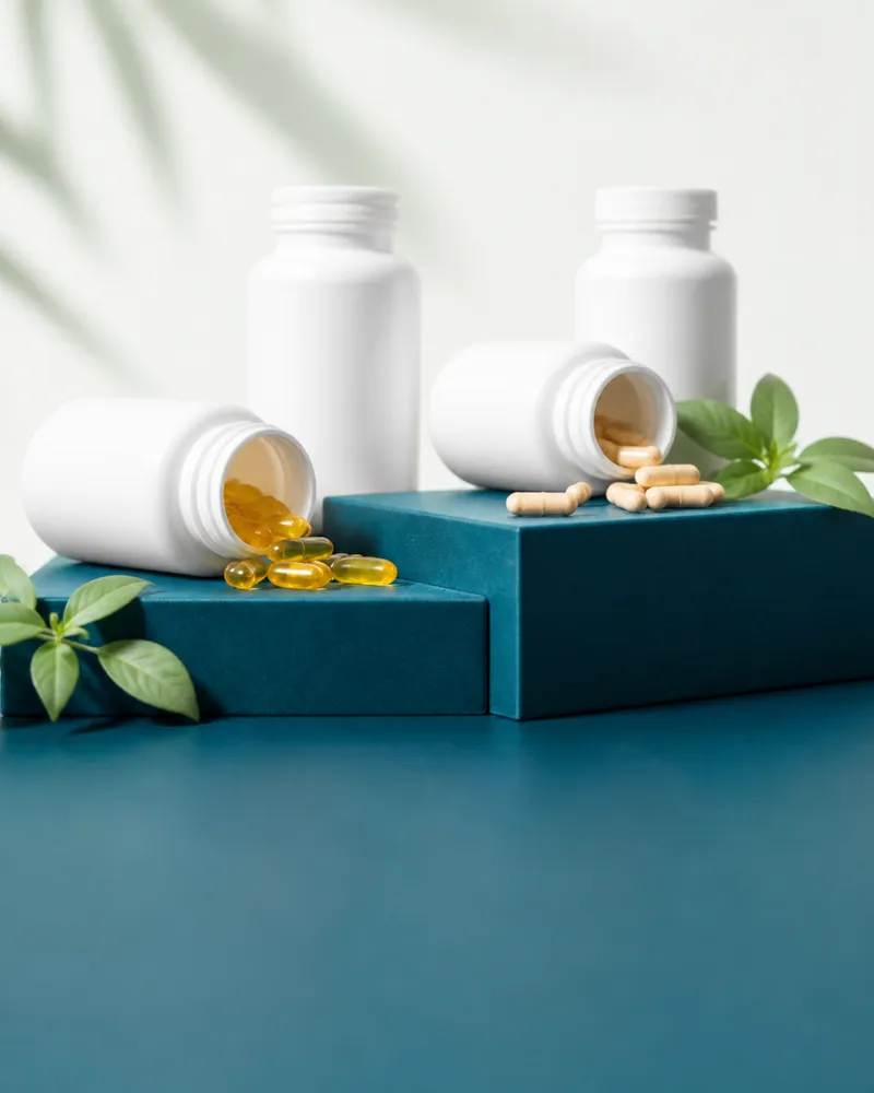 White supplement bottles with softgels and capsules spilling onto teal plinths
